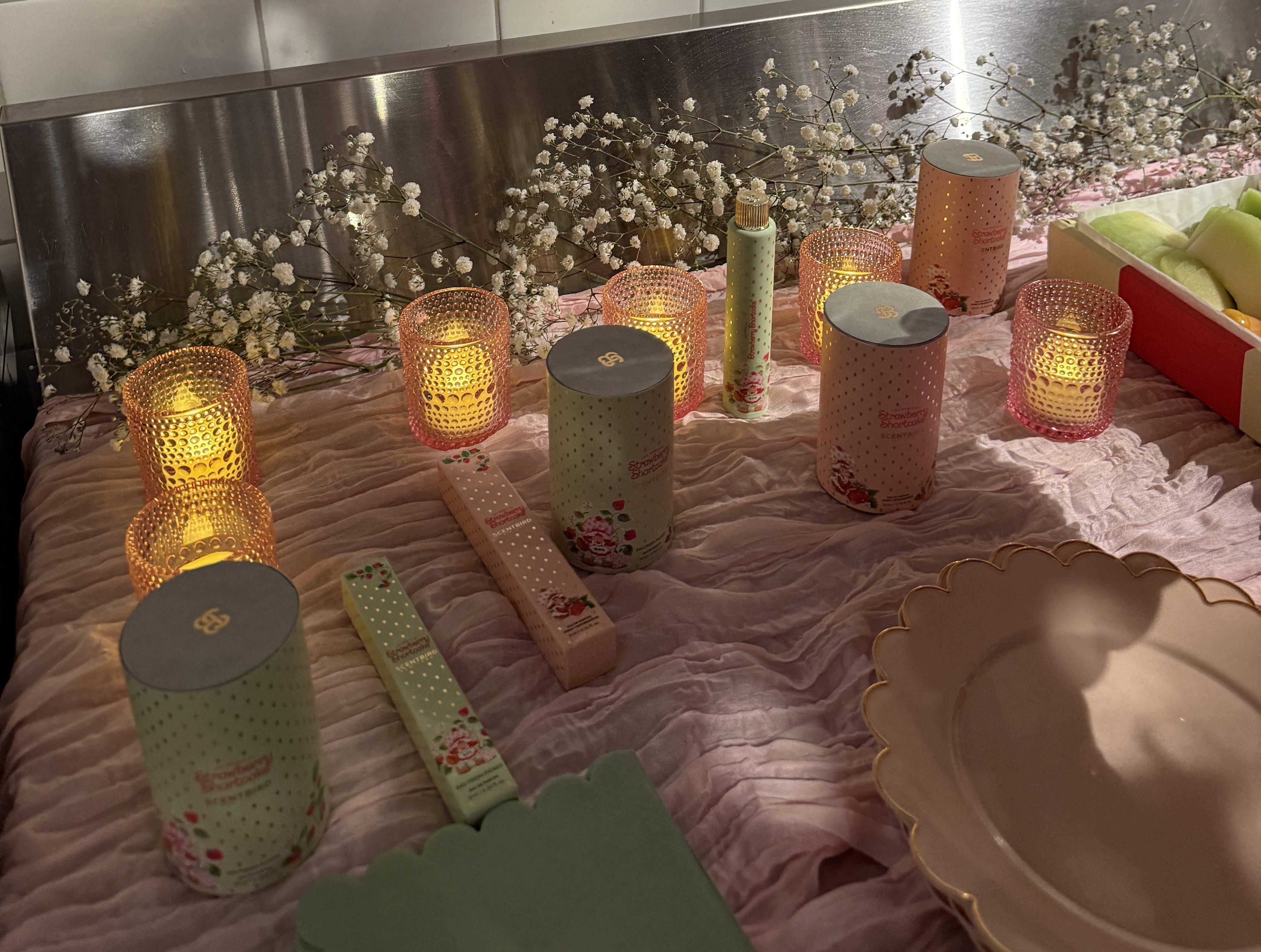 Candles flicker warmly on a pink tablecloth adorned with baby's breath flowers, with Scentbird x Strawberry Shortcake products arranged elegantly beside soft plates and pastel packaging, creating a cozy, inviting atmosphere.