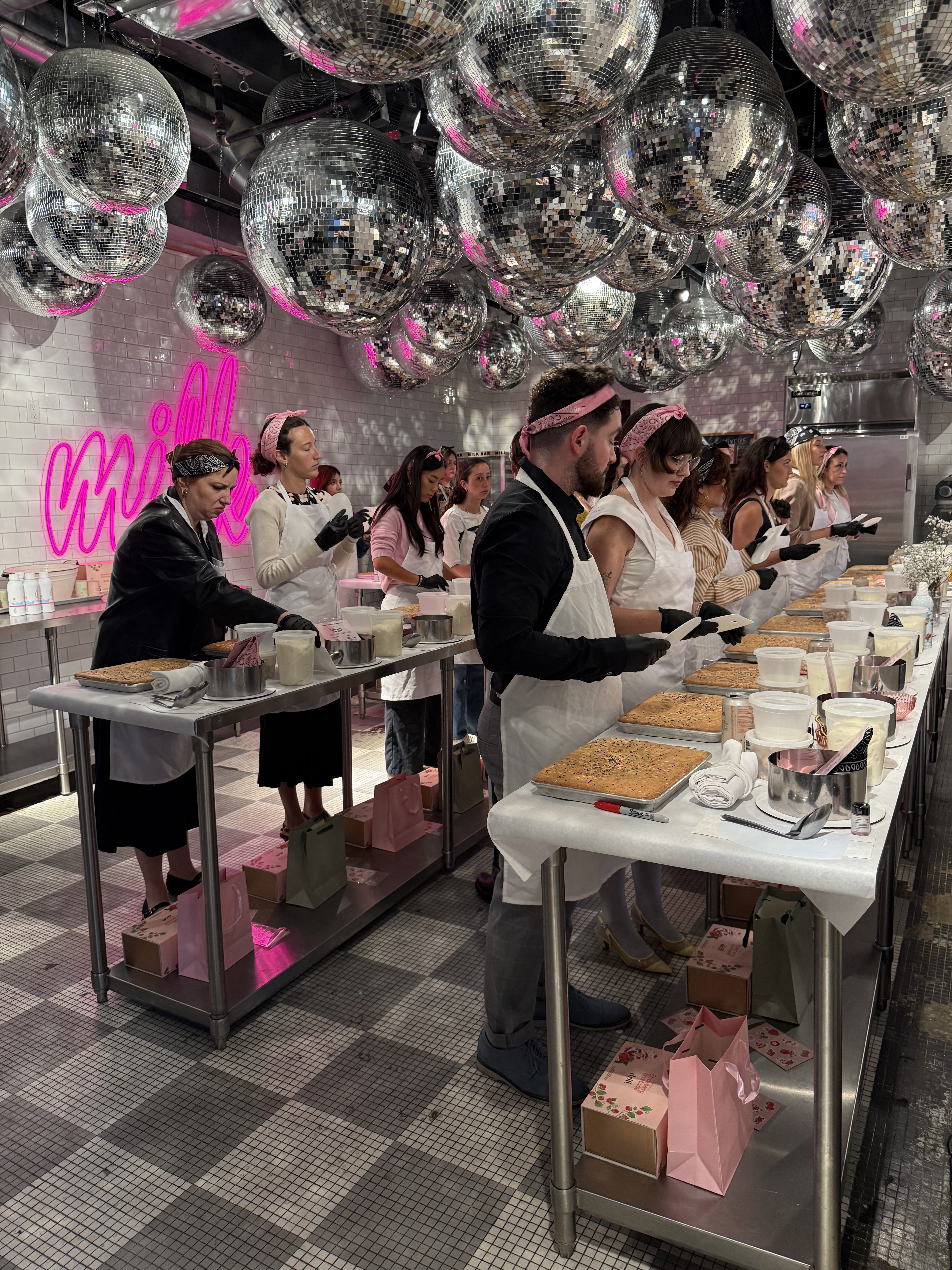 People wearing aprons and gloves are gathered around tables preparing desserts in a bakery with disco balls on the ceiling and a neon pink "milk" sign, which is the Milk Bar logo.