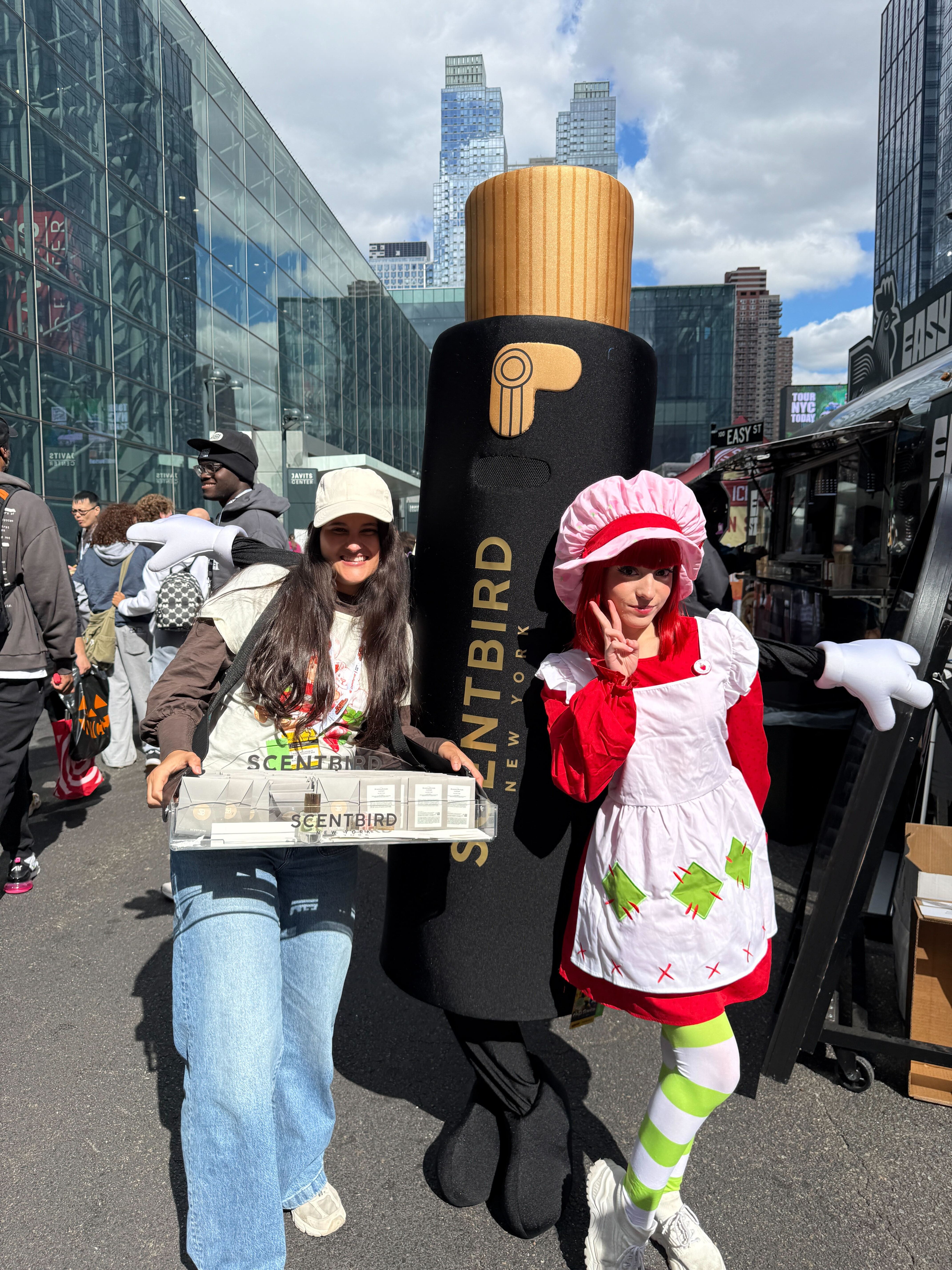 A woman in a Scentbird T-shirt holds a box with samples, standing next to the Scentbird mascot, Misty, and a woman in a Strawberry Shortcake costume at Comic Con in New York City.