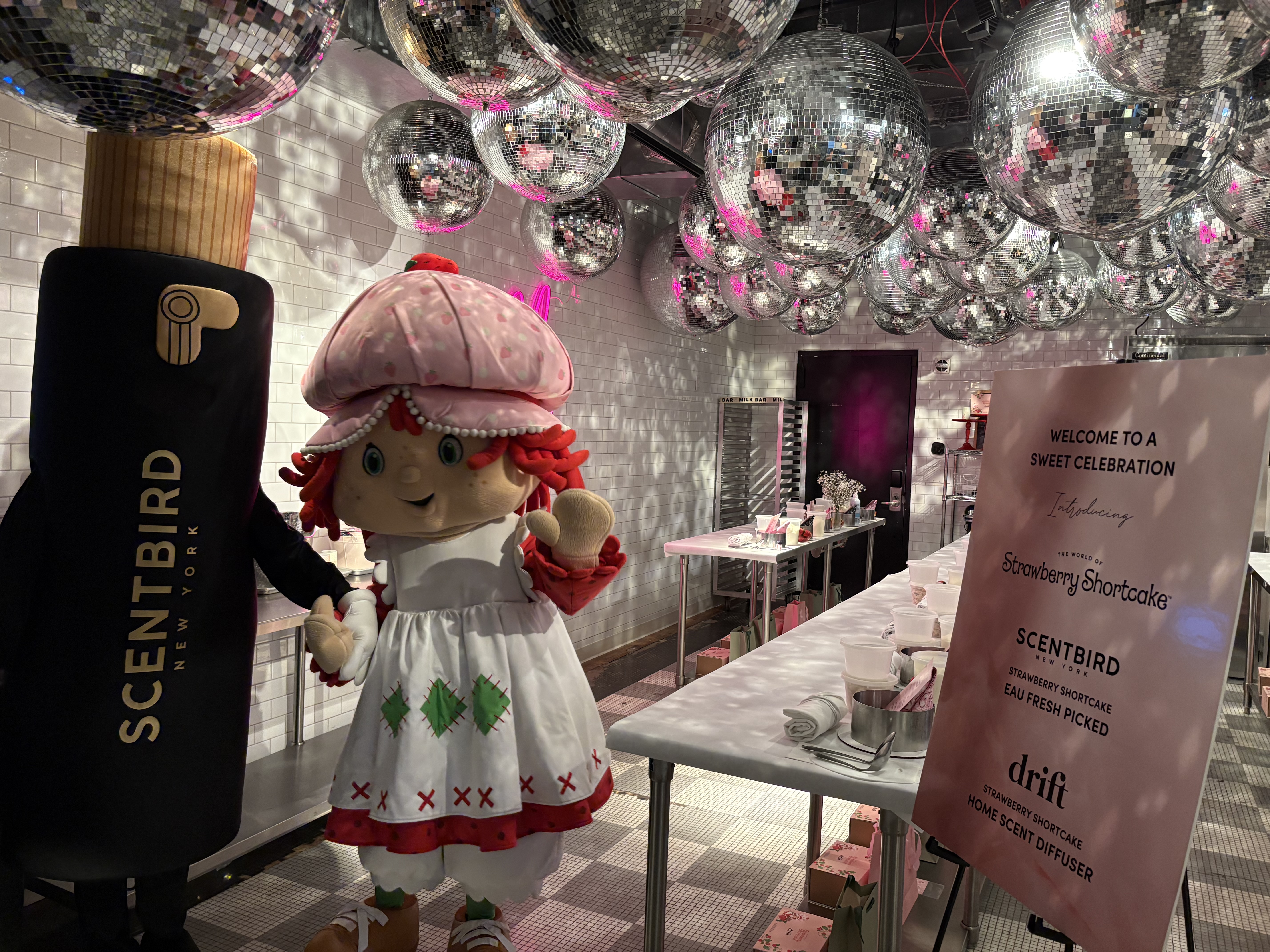 A festive scene with a person in a Strawberry Shortcake costume beside a giant Scentbird product mascot, Misty. Disco balls hang from the ceiling. A sign reads "Welcome to a Sweet Celebration." The setting gives a joyful and whimsical atmosphere.