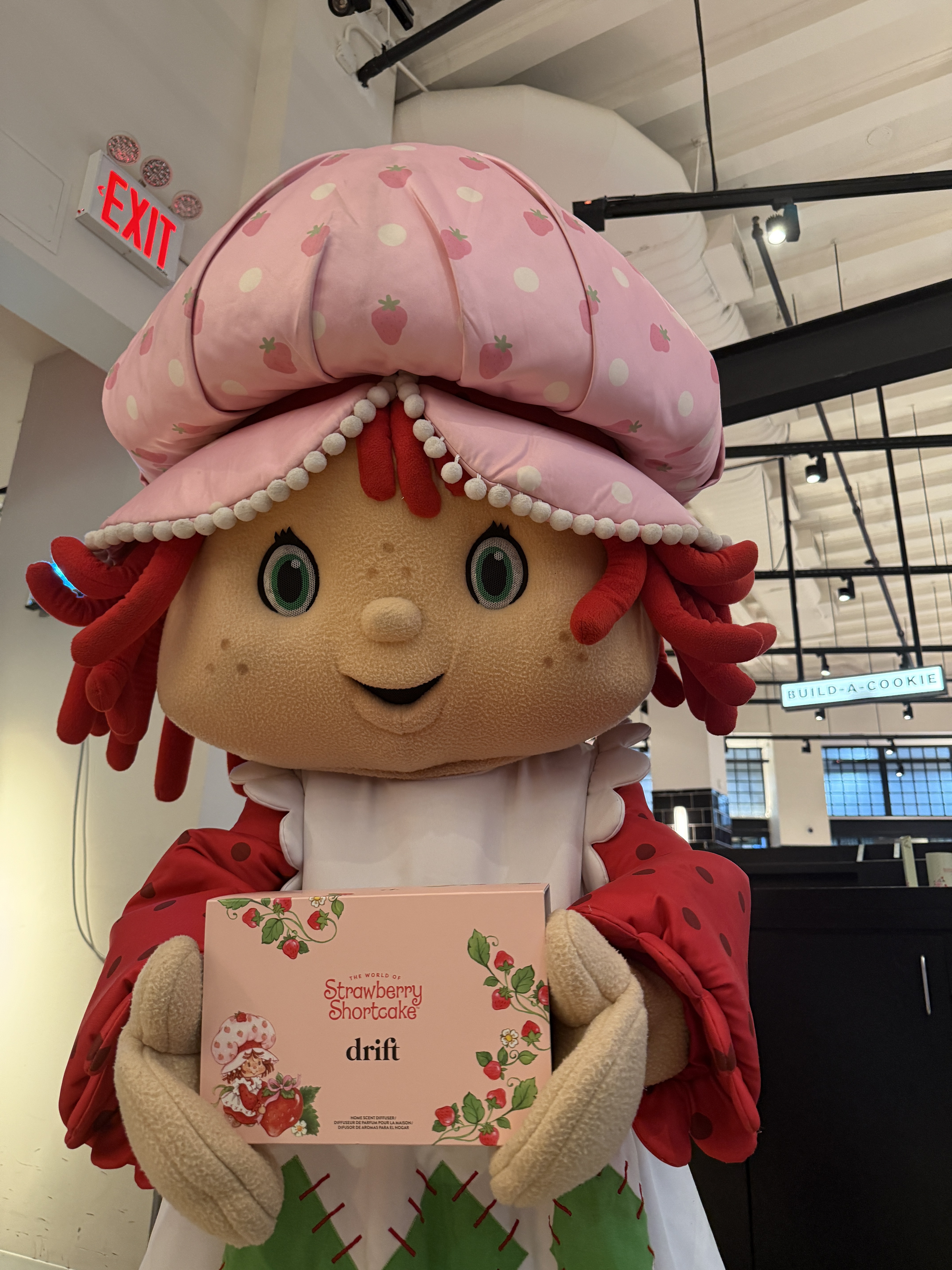 Person wearing a Strawberry Shortcake costume with red hair and a pink strawberry hat holds the Drift x Strawberry Shortcake fragrance gift set box inside a store, smiling and posing for a photo.