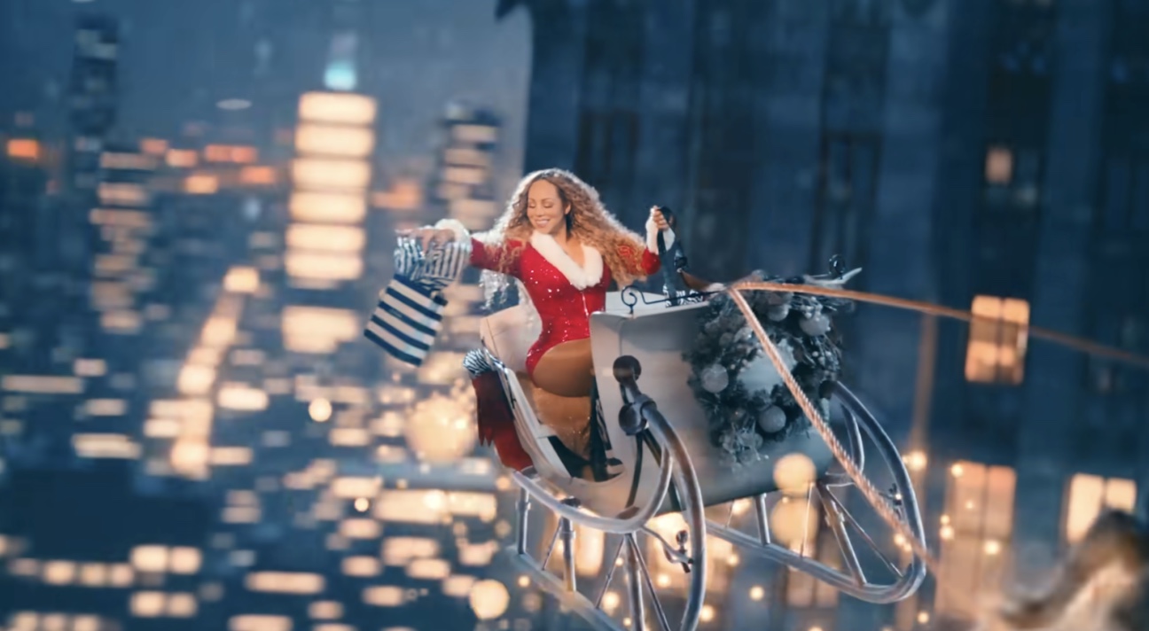Mariah Carey rides a silver sleigh above a city at night, wearing a sparkling red outfit and holding a Sephora bag amid glowing lights.