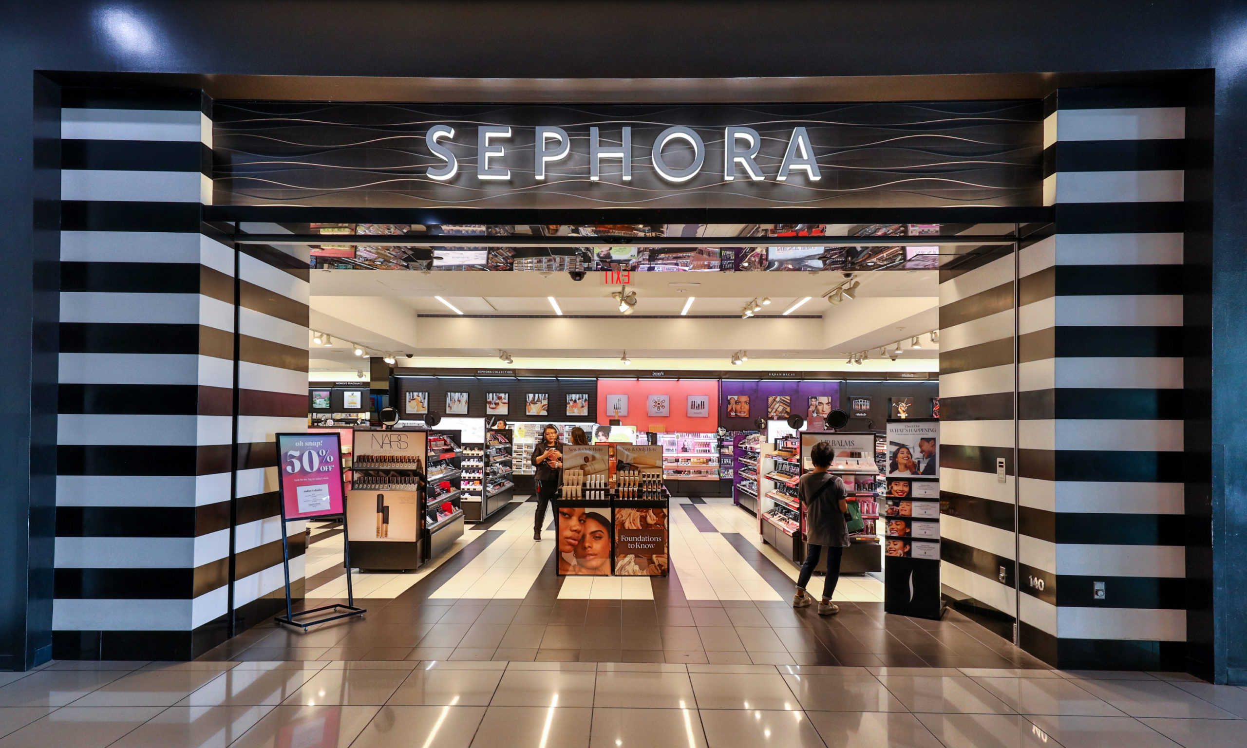 A Sephora store entrance in a mall with black-and-white stripes, a glowing sign, and displays promoting a 50% off sale inside.