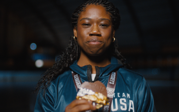 A Team USA athlete wearing a medal and a teal Team USA jacket holds a chocolate ice cream sandwich and smiles at the camera against a dark background.